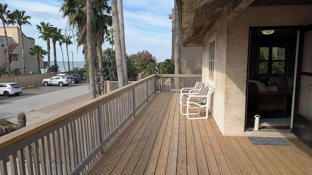 Una terraza de madera con baranda, una puerta que da a una habitación con cama y una silla de mimbre, y palmeras al fondo.