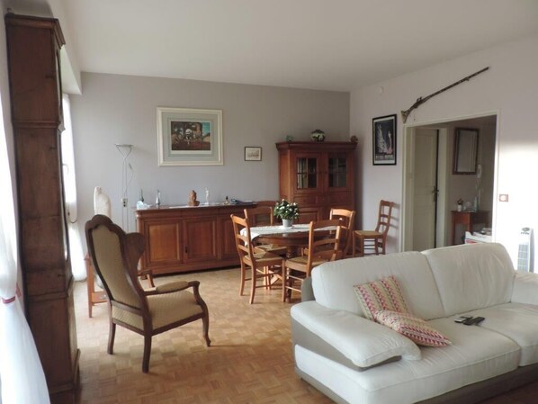 Una sala de estar con un sofá blanco, una mesa de comedor de madera y sillas. Hay un aparador de madera y un cuadro en la pared.