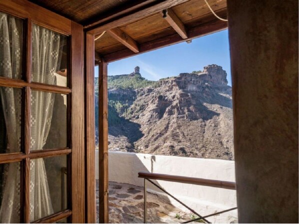 Quarto com teto de madeira, janela com cortinas estampadas e vista para uma paisagem montanhosa rochosa.
