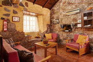 Uma sala de estar rústica com paredes de pedra, teto de madeira, lareira, sofá, duas poltronas e uma mesa de centro.