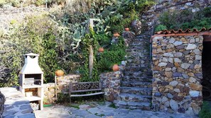Uma escadaria de pedra leva a um edifício rústico de pedra, cercado por vegetação exuberante e uma área de churrasco ao ar livre.