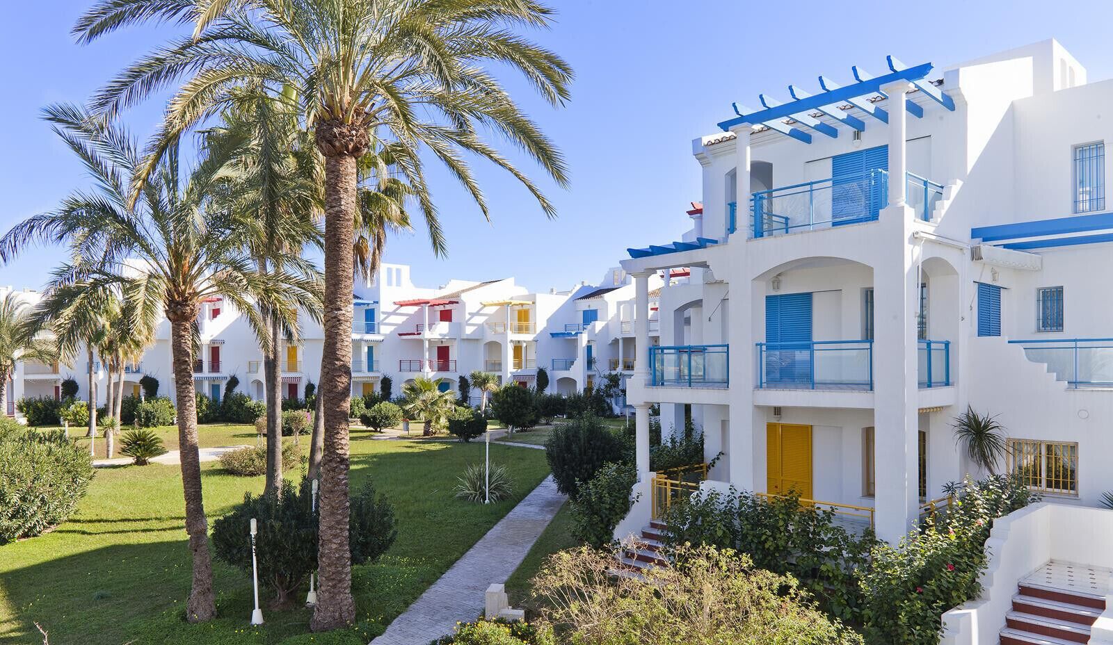 Un complexe hôtelier avec des bâtiments blancs, des balcons bleus et des palmiers.