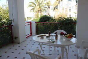 Uma mesa redonda branca com cadeiras, uma tigela de frutas, uma garrafa e um prato com comida em uma varanda com vista para palmeiras e edifícios.