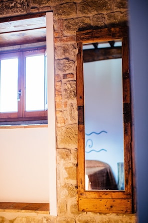 Une pièce rustique avec un mur en pierre, un miroir à cadre en bois et une fenêtre également dotée d’un cadre en bois.