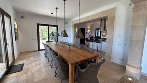 Un comedor amplio con una mesa de madera larga y sillas de mimbre, una cocina al fondo y vistas al verdor a través de la puerta.