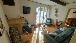 A living room with a sofa, armchair, and a small round table. There is a television mounted on the wall and a window with a view of trees. - Charming cottage in the countryside between St Rémy de Provence and Avignon (Eyragues)