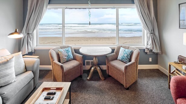 Ein gemütliches Zimmer mit großem Fenster und Blick auf einen Strand, ausgestattet mit zwei Sesseln, einem kleinen Tisch und einem Sofa.