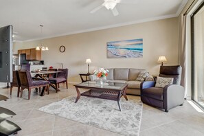 A modern living room with a dining area, a kitchen in the background, and a large window with a view of the beach.
