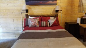 Una habitación con paneles de madera, una cama, dos lámparas fijadas a la pared, un cuadro de montañas y una pequeña mesa con una lámpara.