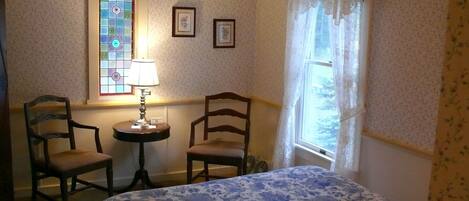 Une pièce comprenant un lit, deux chaises, une table avec une lampe, une fenêtre ornée de vitraux et un papier peint à motifs floraux.