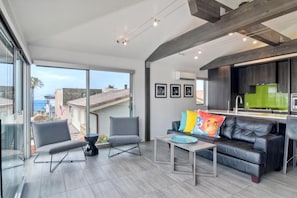 A modern living room with a black leather sofa, two grey armchairs, and a coffee table. A large window offers a view of the cityscape. - Sand (1 bedroom / 1 bath) (Carlsbad)