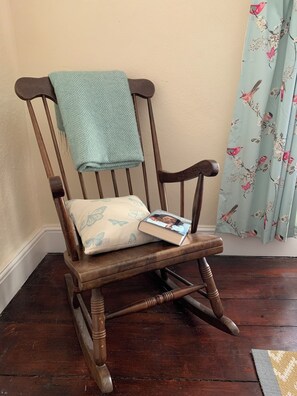 A wooden rocking chair with a cushion and a blanket, positioned against a wall with a floral curtain. - Family-friendly cottage - walk to stunning beach (Angle)