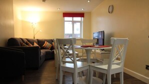 Un salon comprenant un canapé, une table à manger avec des chaises, un téléviseur et une fenêtre agrémentée d’un lambrequin rouge.