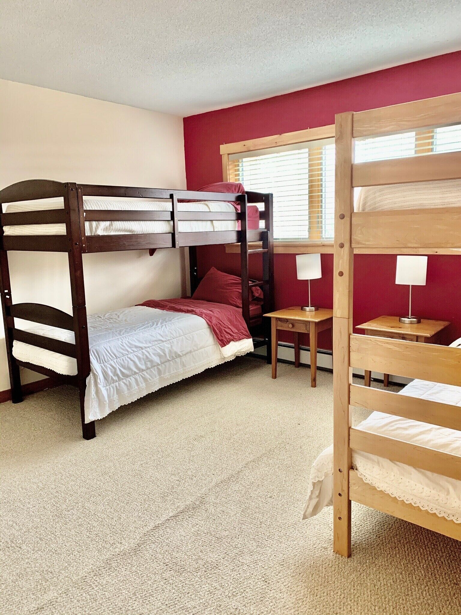 A bunk bed room with a red wall, a window with blinds, and a small table with a lamp.