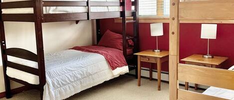 A bunk bed room with a red wall, a window with blinds, and a small table with a lamp.