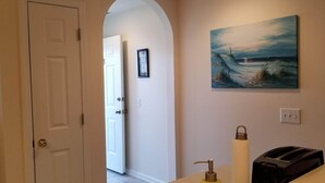 Um corredor com uma porta branca, um quadro na parede, um frasco de álcool em gel e uma torradeira em um balcão.