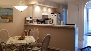 Uma sala de jantar com uma mesa de vidro, cadeiras de vime e uma cesta de frutas. Uma cozinha com armários e eletrodomésticos brancos é visível ao fundo.