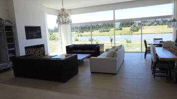 A spacious living room with a large window offering a view of a lake and greenery. There is a black sofa, a white sofa, and a dining table with chairs.