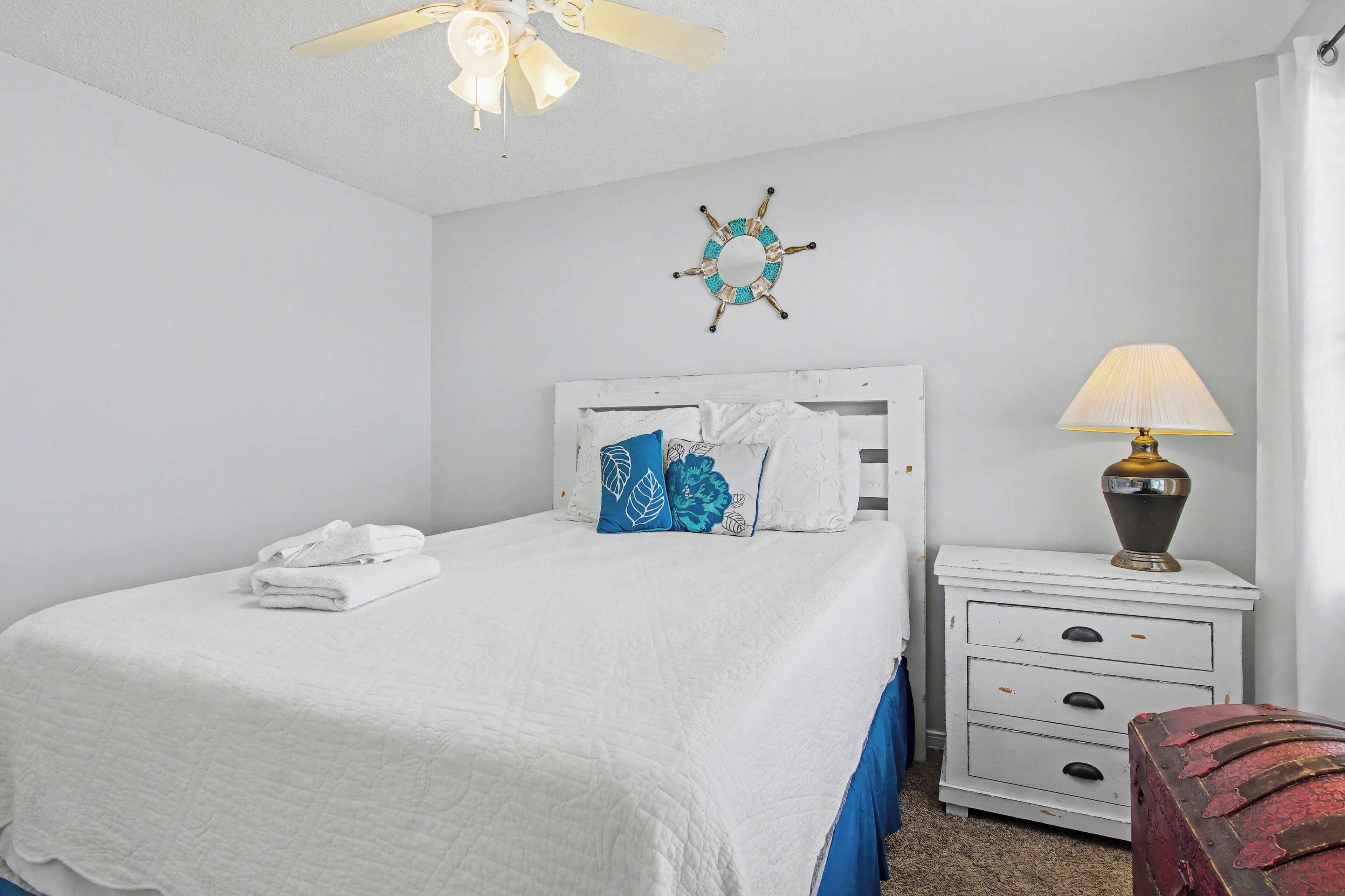 A neatly made bed with white bedding and blue pillows, a white nightstand with a lamp, and a wall decoration.