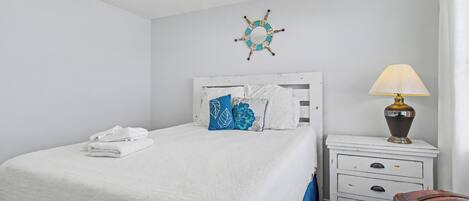 A neatly made bed with white bedding and blue pillows, a white nightstand with a lamp, and a wall decoration.