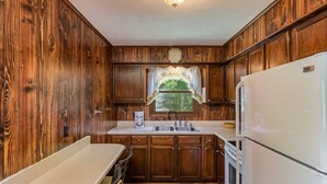 A compact kitchen with wooden cabinets, a sink, a refrigerator, and a window with a floral curtain.