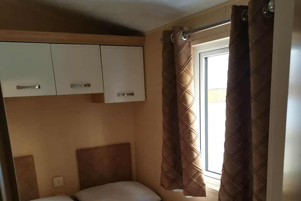 A single bed with a headboard, a window with curtains, and a wardrobe with two drawers.