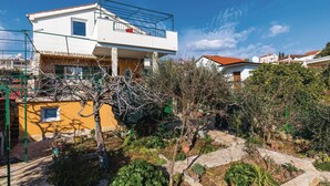A residential area with a white two-story house, a yellow building, and a garden with various plants and a stone pathway. - Awesome apartment in Seget Vranjica with WiFi (Seget Vranjica)