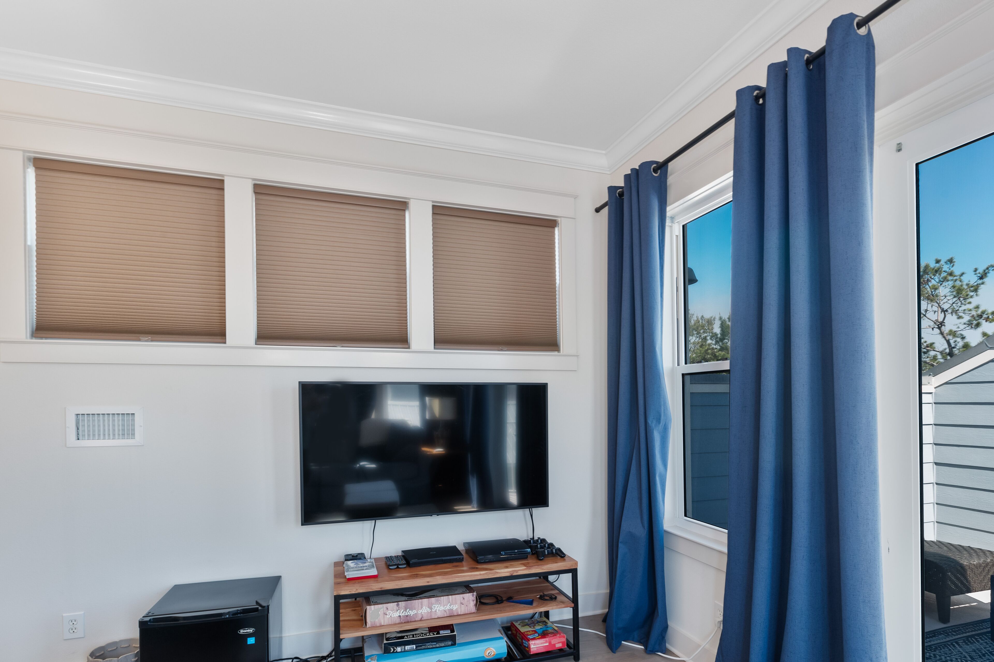 A room with a flat-screen TV mounted on the wall, a wooden entertainment unit with electronic devices, and a window with blue curtains.