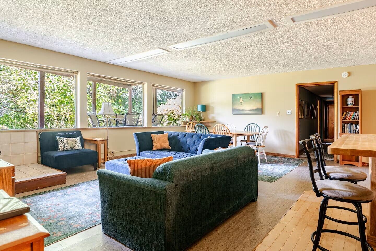 Ein geräumiges Wohnzimmer mit einer großen Couch, einem Esstisch und mehreren Stühlen. Große Fenster sorgen für natürliches Licht und einen Blick ins Grüne.