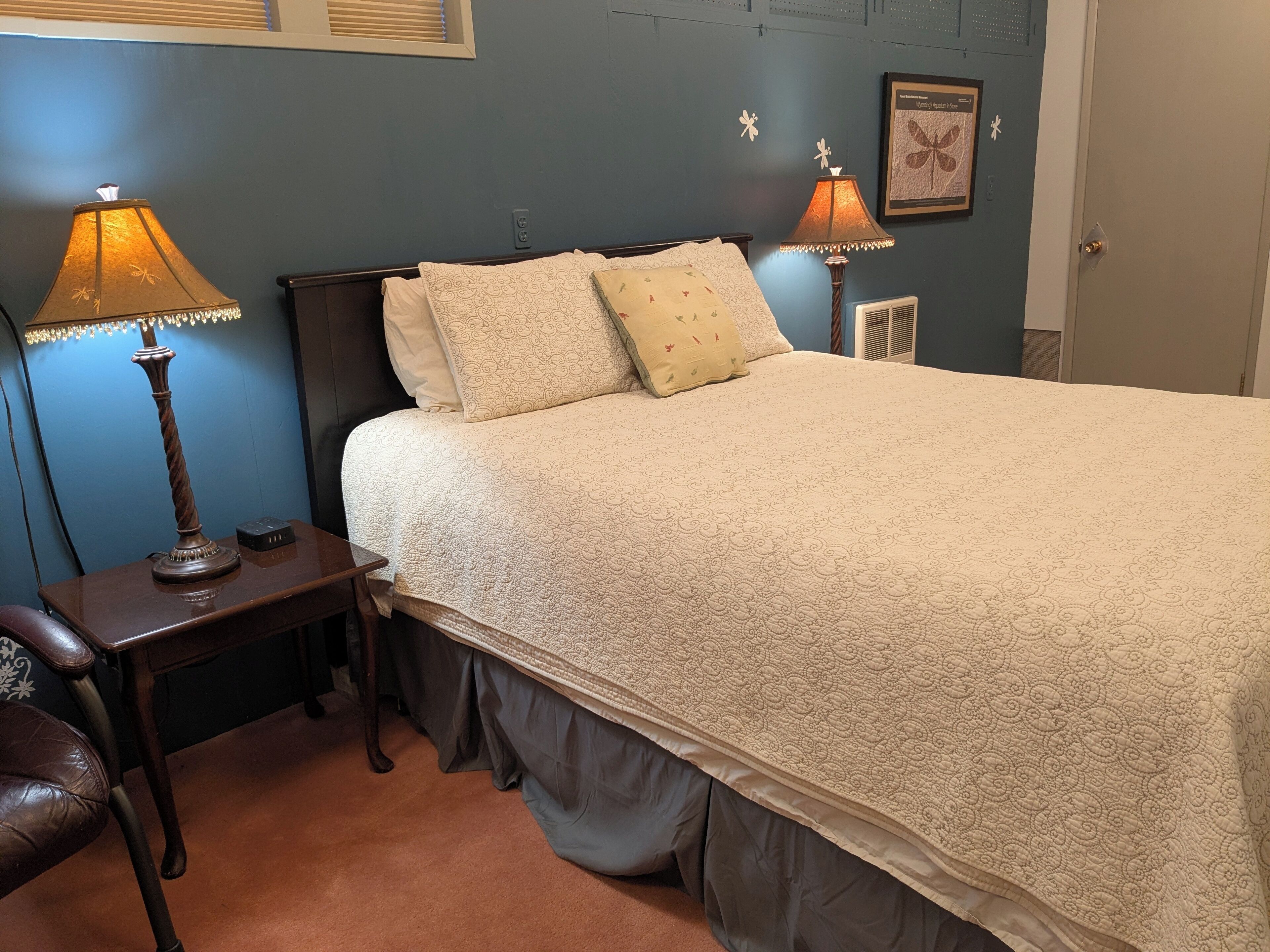 A bedroom with a bed, two bedside lamps, a nightstand, a chair, and a radiator.