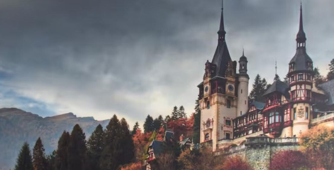 A castle with multiple towers and a prominent clock tower, surrounded by autumn foliage and mountains in the background.