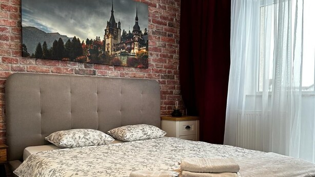 A modern bedroom with a brick wall, a large bed with floral bedding, and a cityscape painting above it. - Best Apartment Suceava (Ipotești)