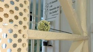 A room with a decorative white partition featuring star-shaped cutouts, a small white flower arrangement on a shelf, and a sign with text in Portuguese.