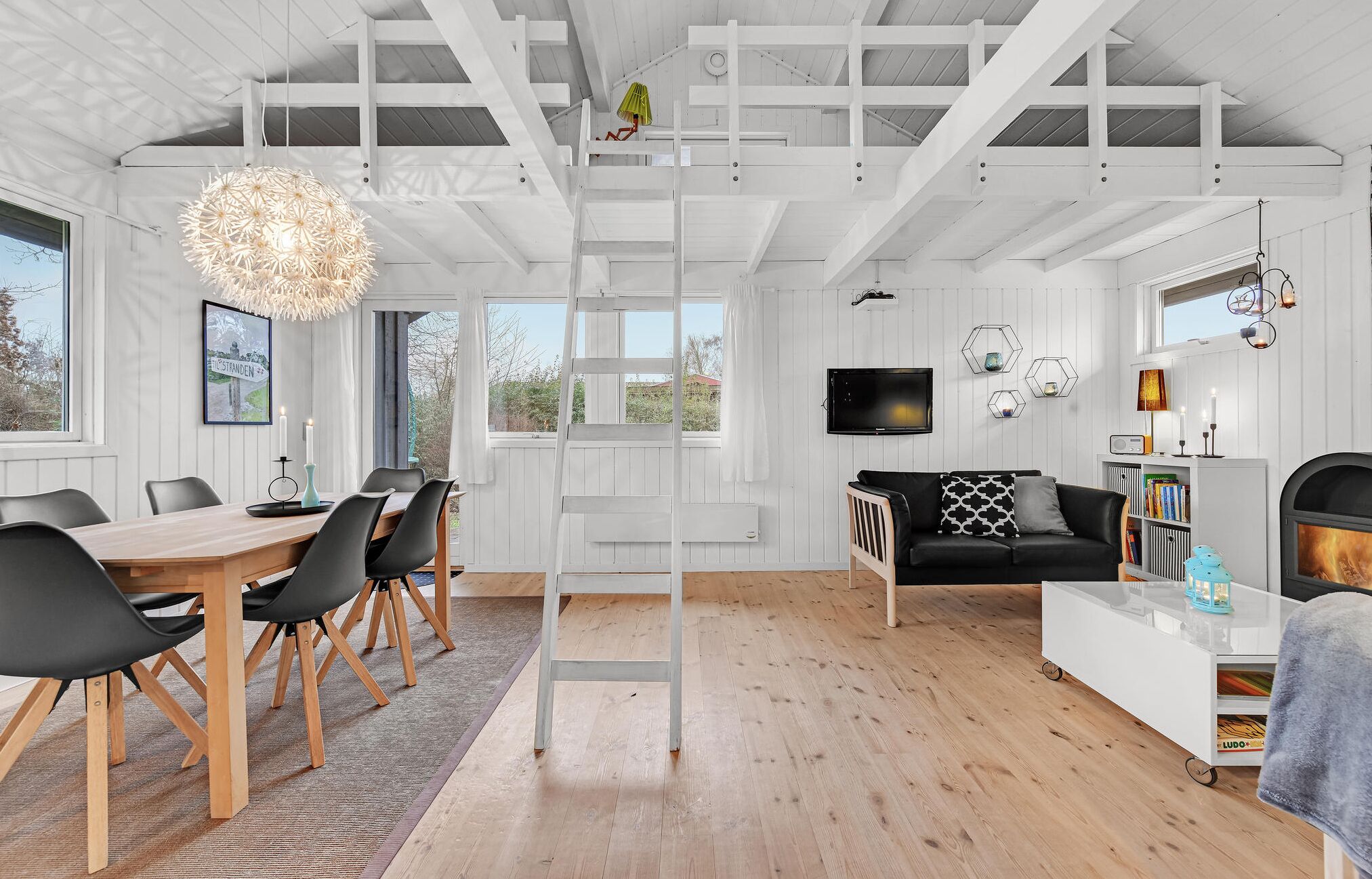 A spacious living room with a high ceiling, a dining area with a wooden table and chairs, a black sofa with patterned cushions, and a fireplace.