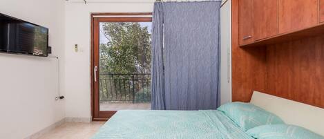 A bedroom with a bed, a TV mounted on the wall, a wooden cabinet, and a balcony with a view of trees.