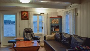 Un salon chaleureux avec un canapé en cuir, une table basse en bois et un fauteuil. On y trouve une bibliothèque remplie de livres et d’objets décoratifs, un lampadaire et, par les fenêtres, une vue sur l’eau.