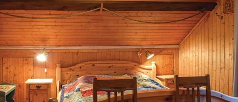 A wooden cabin bedroom with a bed, two chairs, a nightstand, and a lamp.
