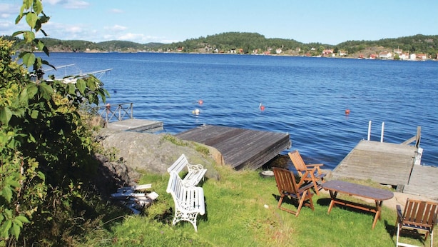 Une jetée au bord du lac, avec des chaises en bois et un banc, une zone herbeuse et une vue sur le lac et les bâtiments situés au loin.