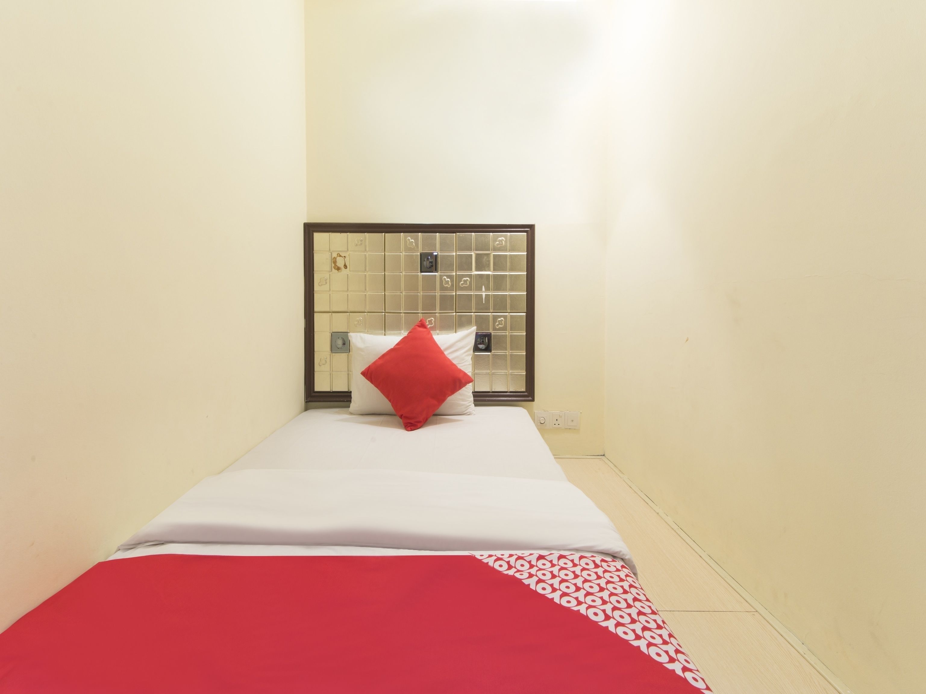 A small, single-bed room with a white bedspread, a red pillow, and a window with a metal grid.