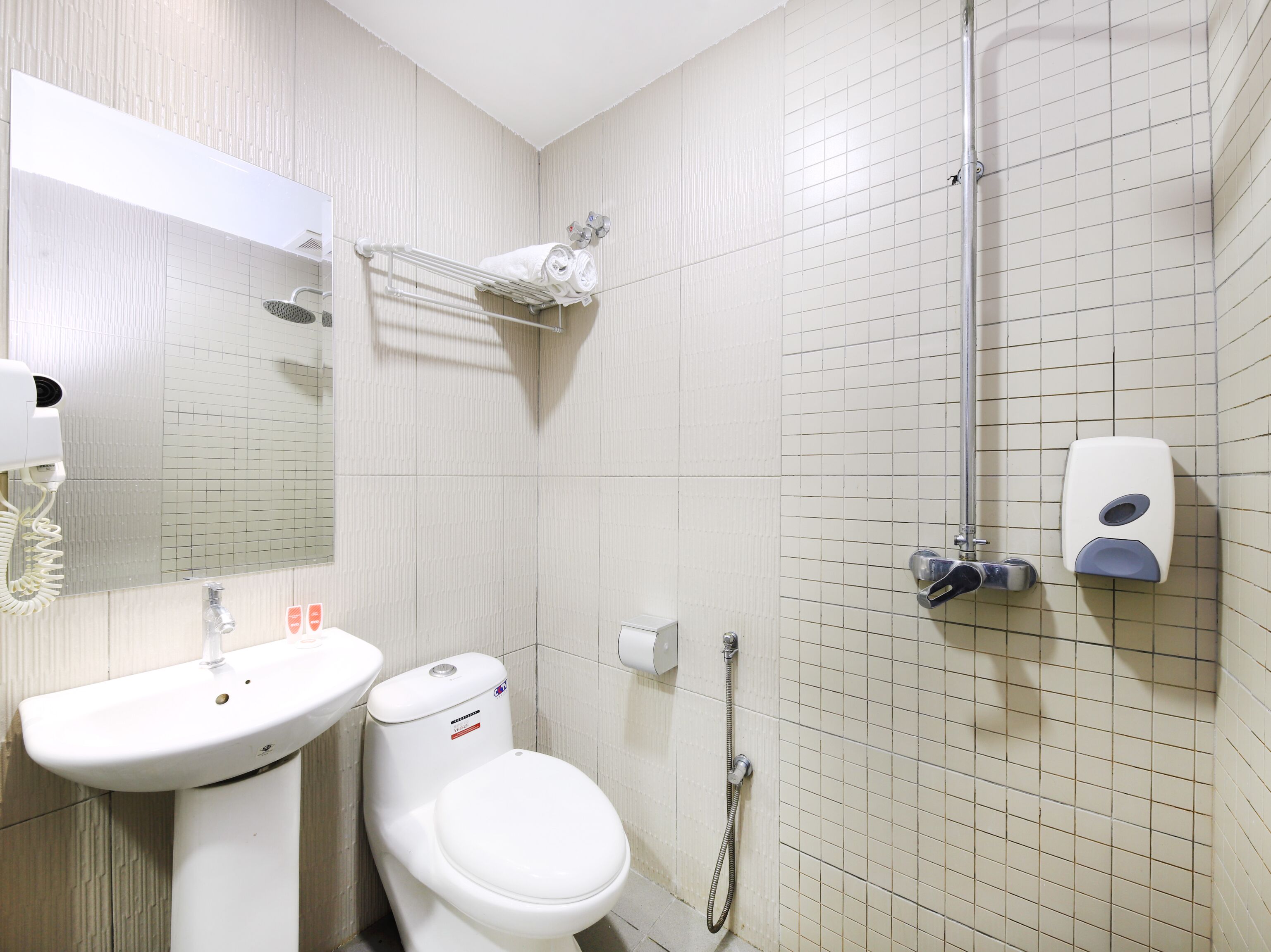 A bathroom with a sink, toilet, mirror, towel rack, shower, and soap dispenser.