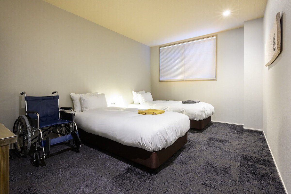 A hotel room with two beds, a wheelchair, a desk, and a window with blinds.