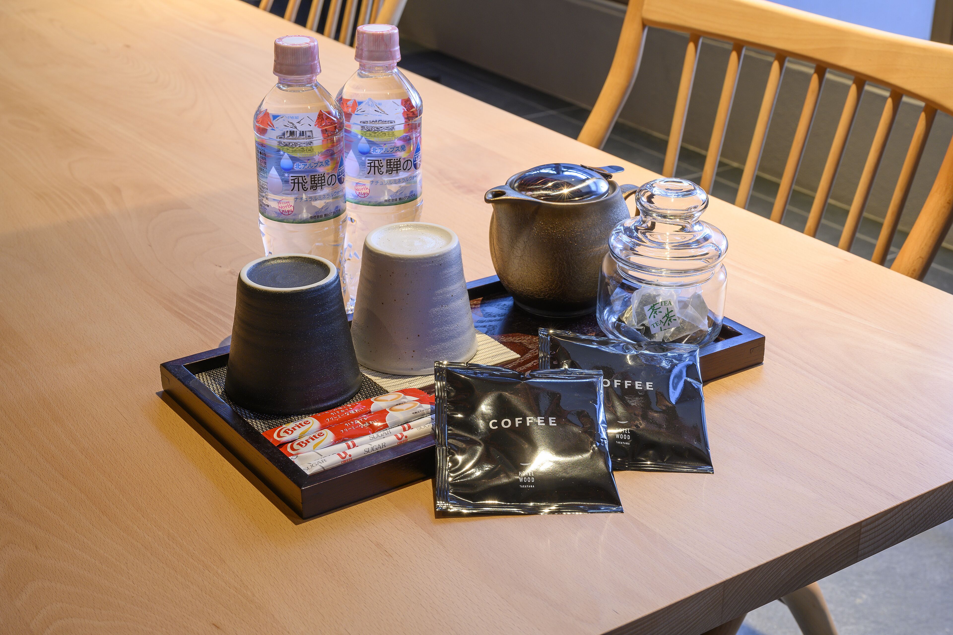 A tray with coffee bags, a teapot, a water bottle, and a mug on a wooden table.