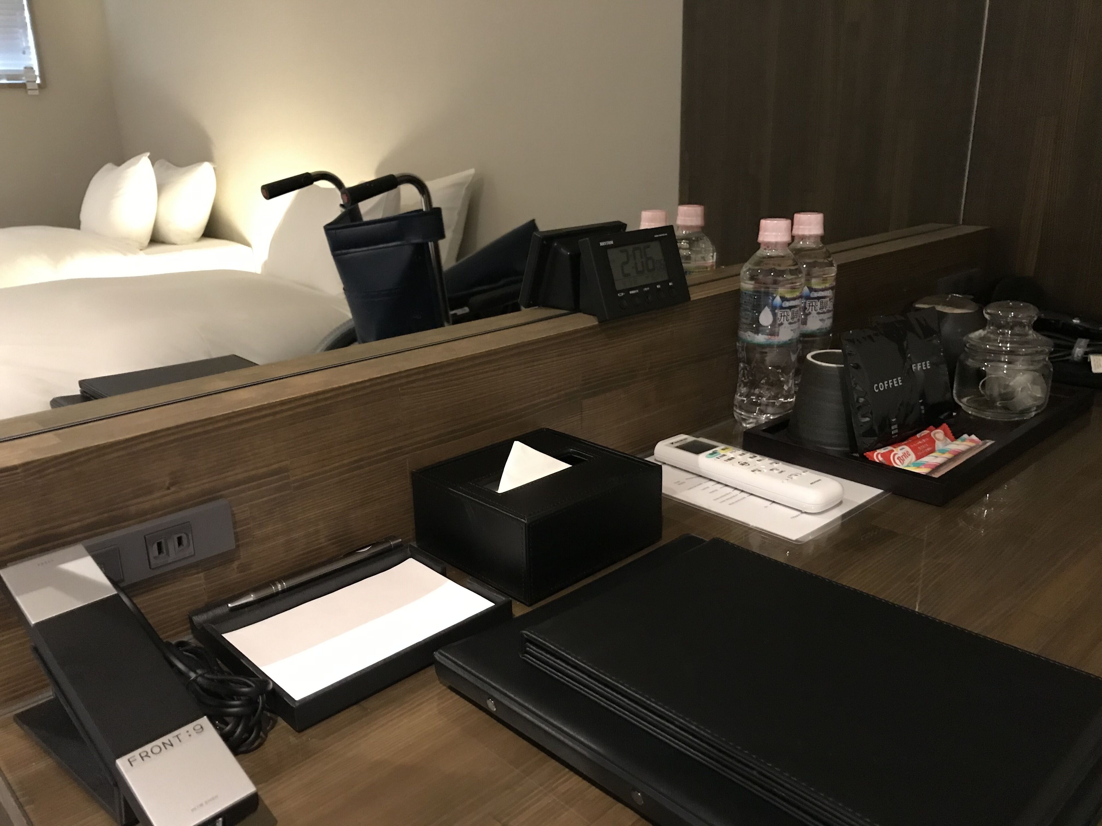 Ein Hotelzimmer mit Holzschreibtisch, einem Bett mit weißen Laken, einer digitalen Uhr, Wasserflaschen, Kaffeebeuteln und einer Fernbedienung.