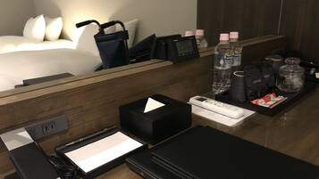 Ein Hotelzimmer mit Holzschreibtisch, einem Bett mit weißen Laken, einer digitalen Uhr, Wasserflaschen, Kaffeebeuteln und einer Fernbedienung.