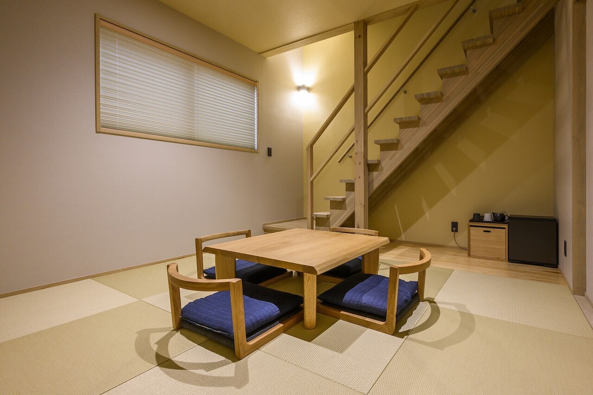 A room with tatami flooring, a wooden table with cushions, a staircase, and a wall-mounted shelf.