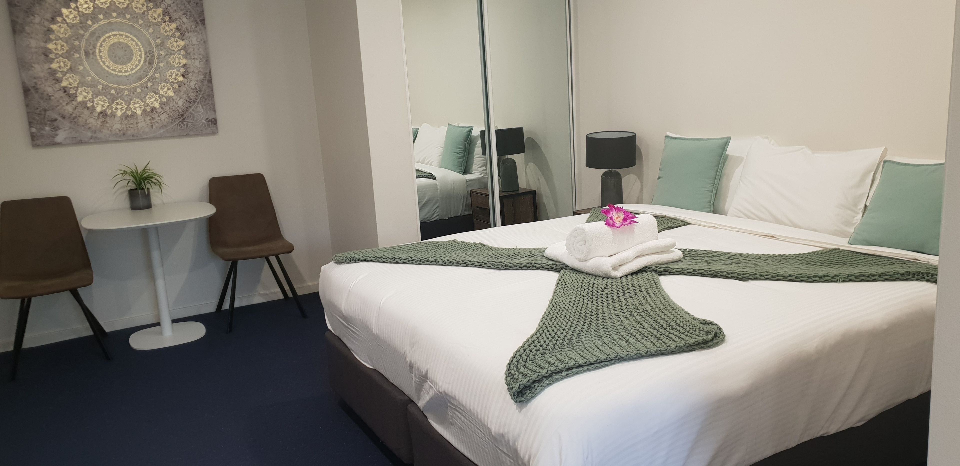 Quarto de hotel com cama, uma mesinha, duas cadeiras, um vaso de planta e um espelho grande.