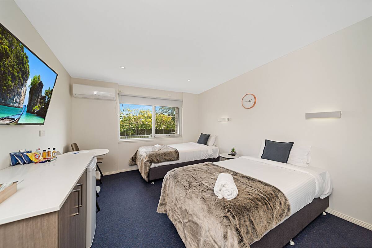 A hotel room with two beds, a desk, a TV, and a window with a view of trees.