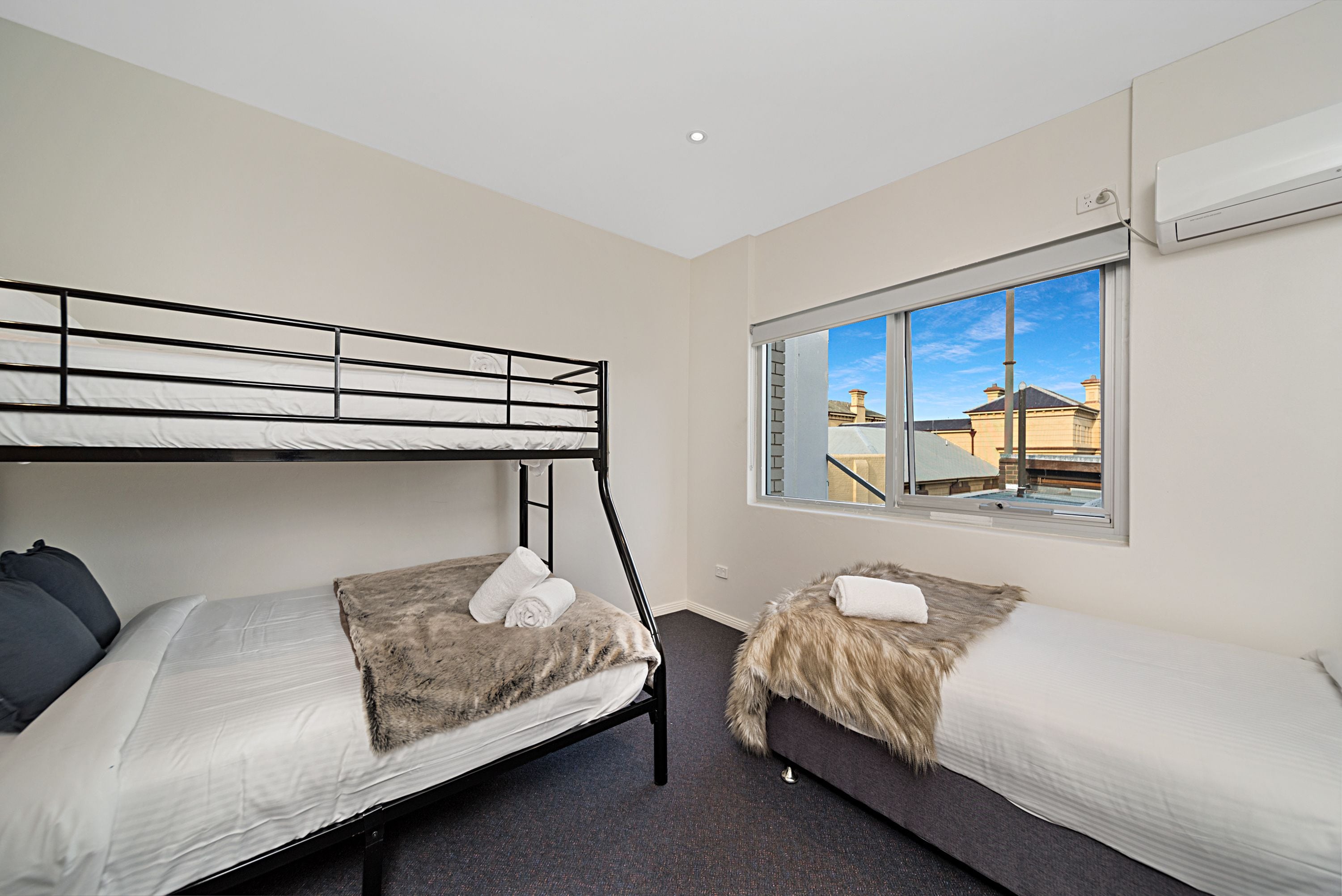 A room with two bunk beds, a window with a view of buildings, and an air conditioning unit on the wall.