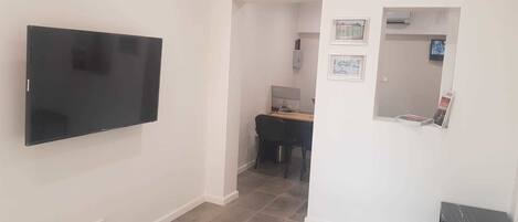 A living room with a flat-screen TV mounted on the wall, a dining area with a table and chairs, and a small black side table.