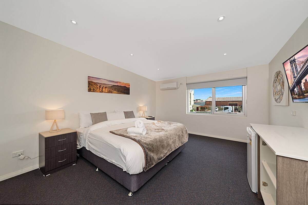 A modern bedroom with a large bed, bedside tables, a window with a city view, and artwork on the walls.